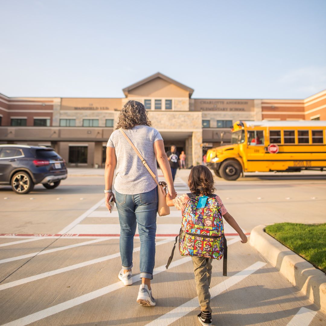 woman walks beside kid wearing backpack towards school building